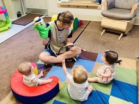 Infant teacher sitting on the floor with three children sitting around her while reading a book with an Og puppet on her hand.