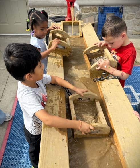 Children outside participating in hands-on outdoor sensory play using wooden sifters to uncover gems in dirt