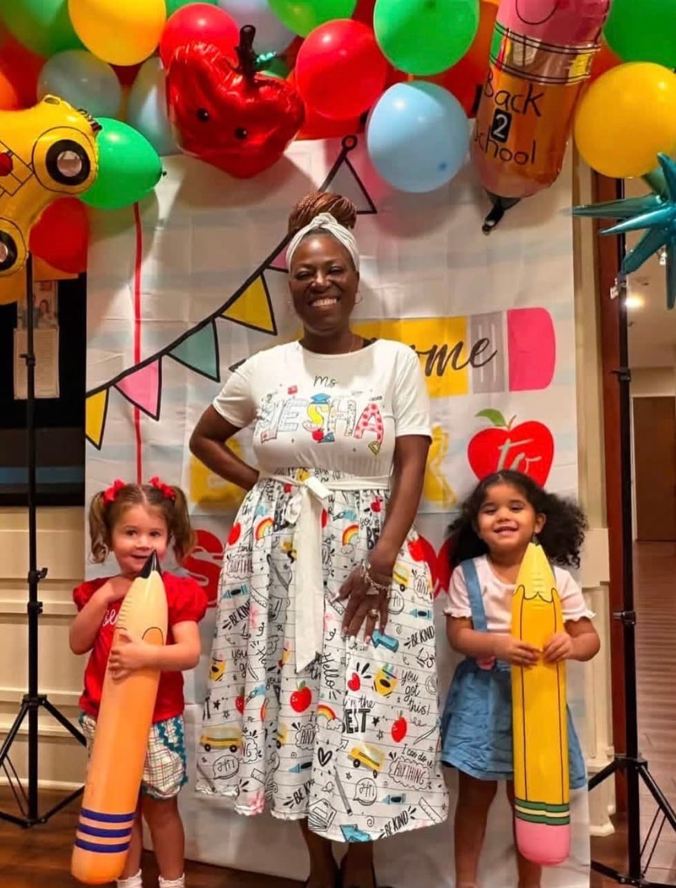 Two children and a teacher pose with First Day of School props and balloons in colorful attire
