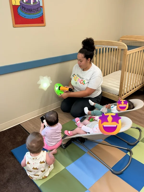 A group of infants gather around as a teacher uses a shape toy and flashlight to project stars on the wall.