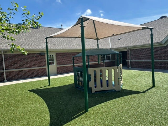 Infant outdoor play area at Primrose School of Eagan featuring an age-appropriate play structure with shade sail coverage, lush artificial turf, and secure fencing adjacent to the school building