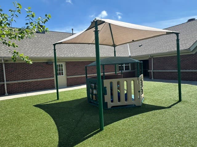 Infant outdoor play area at Primrose School of Eagan featuring an age-appropriate play structure with shade sail coverage, lush artificial turf, and secure fencing adjacent to the school building