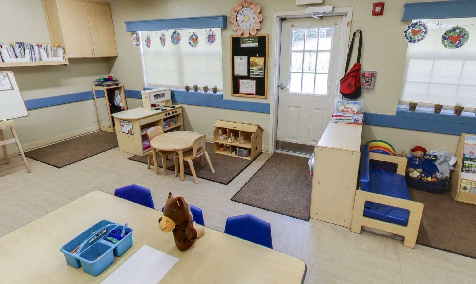 Primrose School of Eagan kindergarten classroom featuring a reading corner with book hospital, dramatic play kitchen, dollhouse, learning table with blue chairs, children's artwork garland, and a Safe School Center board near the door.