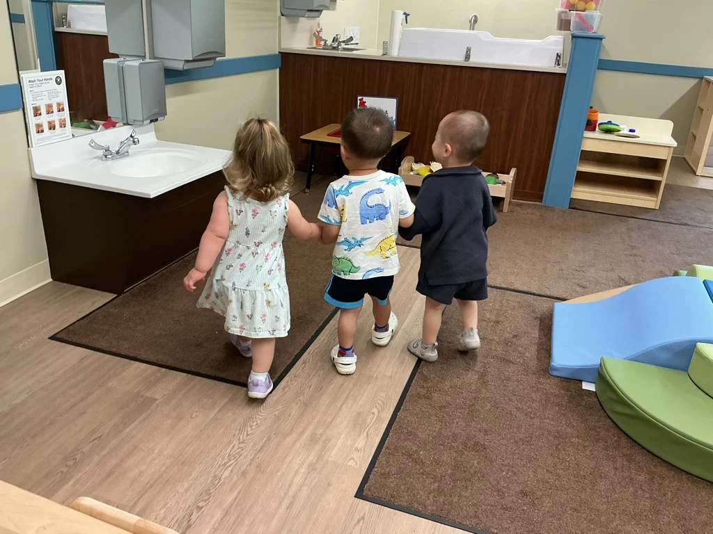 Three toddlers, holding hands as they walk through the classroom.