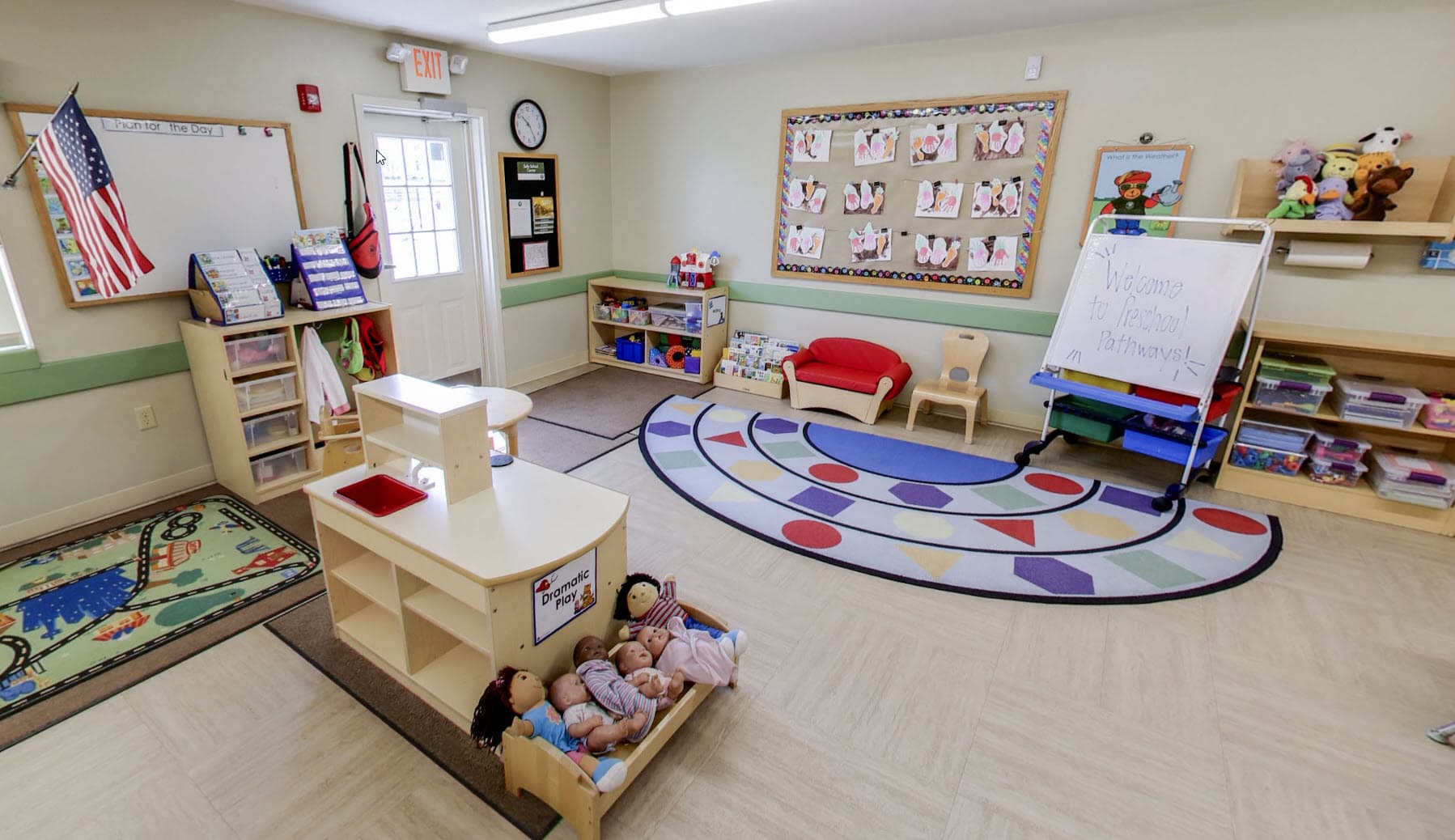 Primrose School of Eagan Preschool Pathways classroom featuring a dramatic play area, colorful shape rug, children's handprint artwork bulletin board, whiteboard with "Welcome to Preschool Pathways," and multiple learning centers.