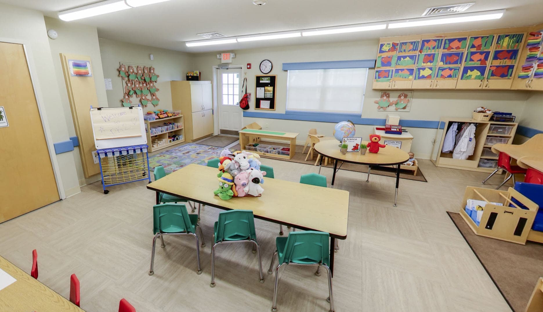 Primrose School of Eagan Pre-Kindergarten classroom featuring group learning tables, a science center with globe, dramatic play area with lab coats, Primrose Friends stuffed animals, and colorful fish artwork displayed across cabinet doors.