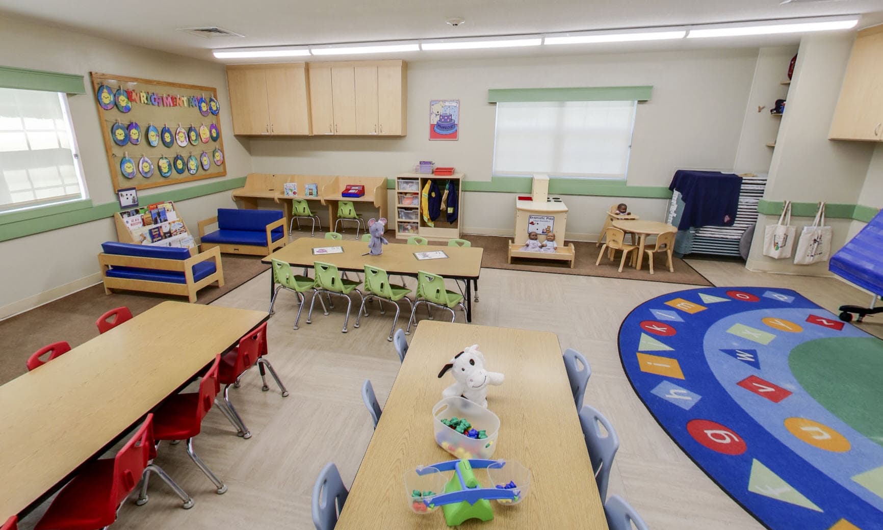 Primrose School of Eagan preschool classroom featuring multiple learning tables, a letters and numbers rug, reading corner with sofa, dramatic play area, and children's artwork displayed on a bulletin board.
