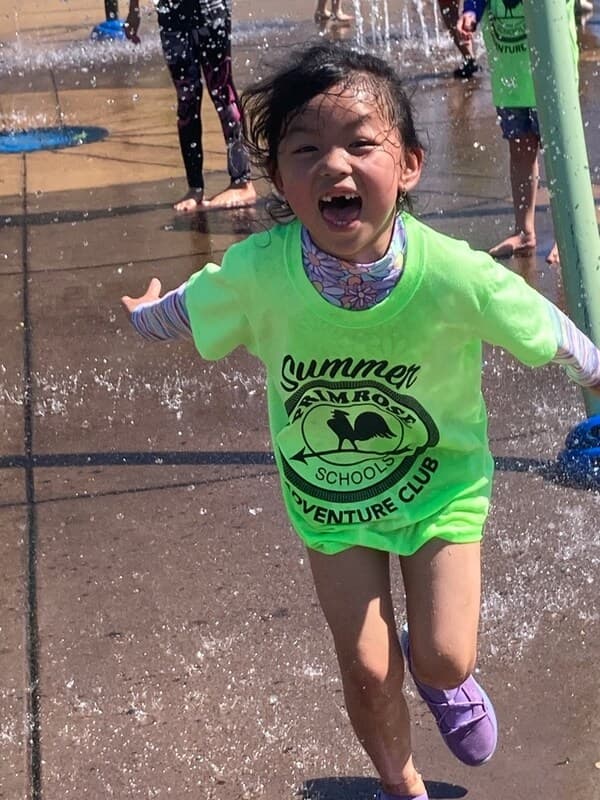 A five year old girl is running through a splash pad, wearing a Primrose Schools t-shirt, with a big smile on her face