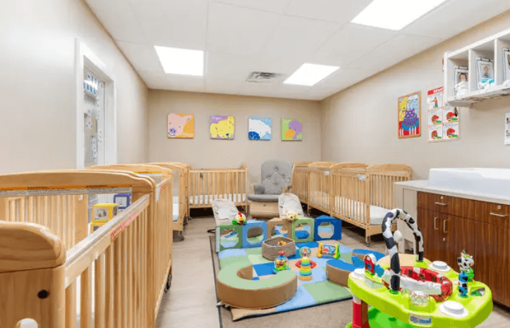 bright, clean classroom with beige walls and a window, wooden cribs, rocking chair, colorful play mat and toys. Paintings of the Primrose friends on the walls