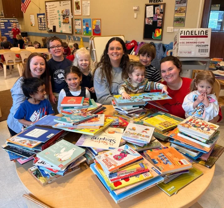 Teachers and students gathered around a classroom table stacked high with donated books at