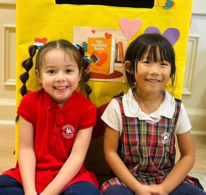 Two preschool students in Primrose uniforms sitting beside a decorated book donation box at Primrose Denver North