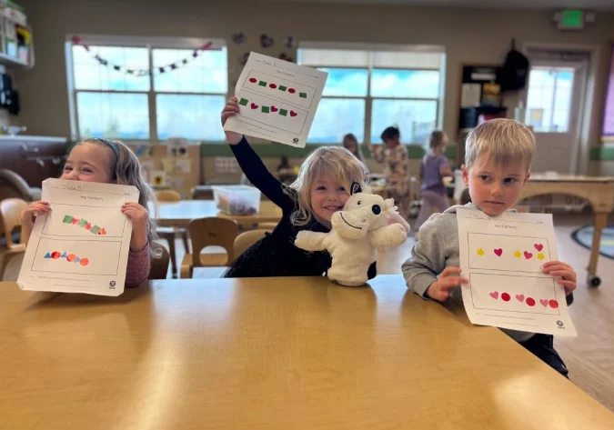Three preschool students holding up completed pattern worksheets in a classroom at Primrose Denver north