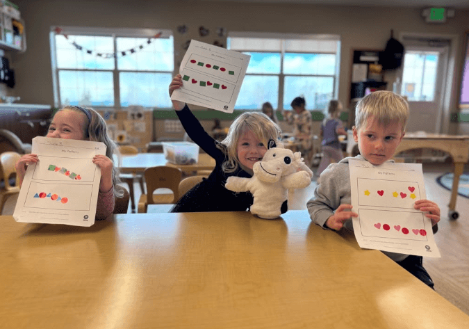 Three preschool students holding up completed pattern worksheets in a classroom at Primrose Denver north