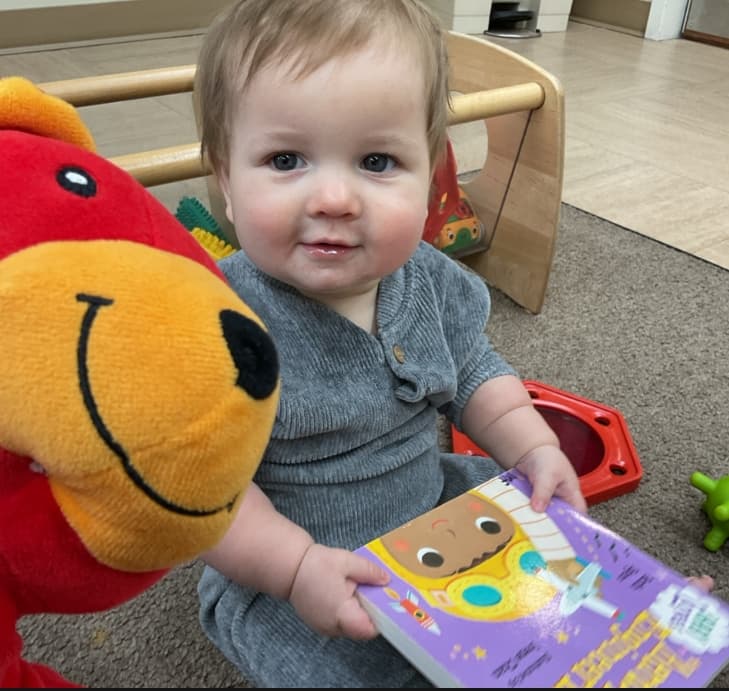 Infant holding a colorful board book while engaging with a Primrose classroom character puppet during literacy enrichment.