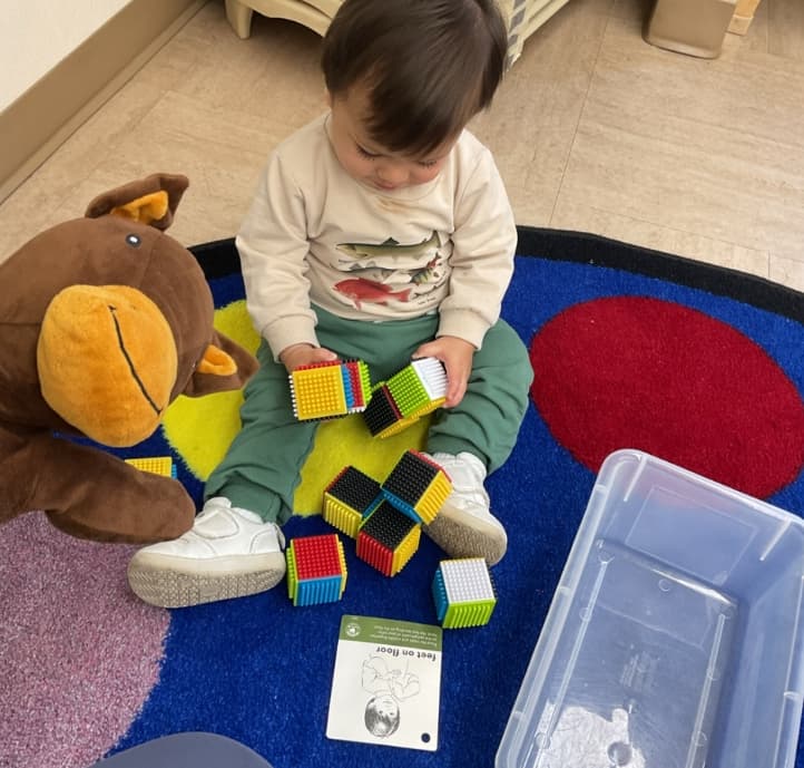 Toddler sitting on a colorful classroom rug exploring sensory building blocks and developing fine motor skills.