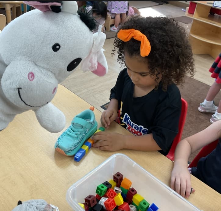 Preschool student using connecting cubes to measure the length of a shoe during a math and problem-solving activity with a classroom puppet nearby.