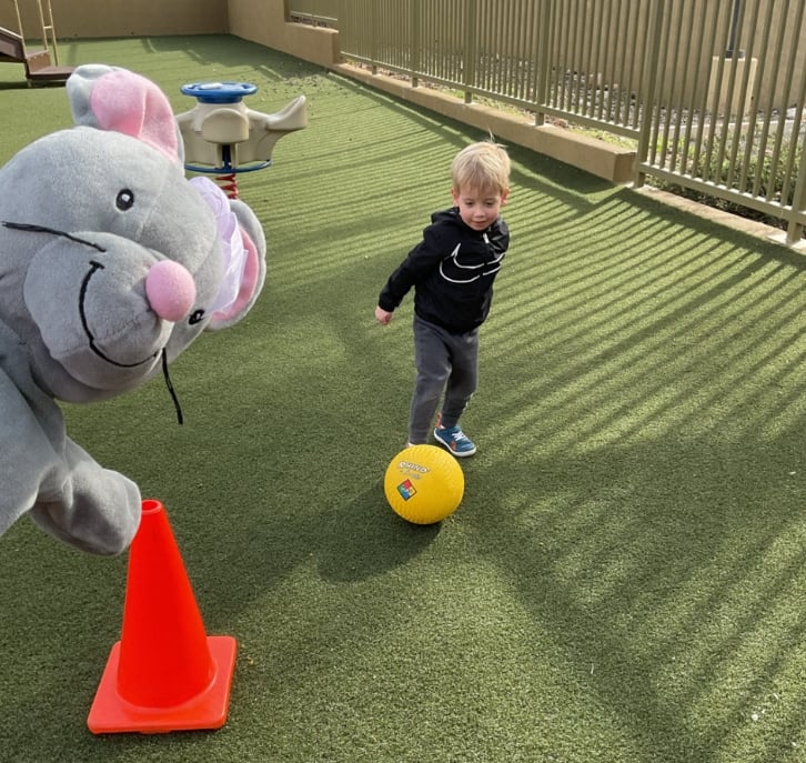 Preschool student practicing gross motor skills by kicking a ball through an outdoor obstacle course while participating in a fun physical activity with a classroom character mascot nearby.