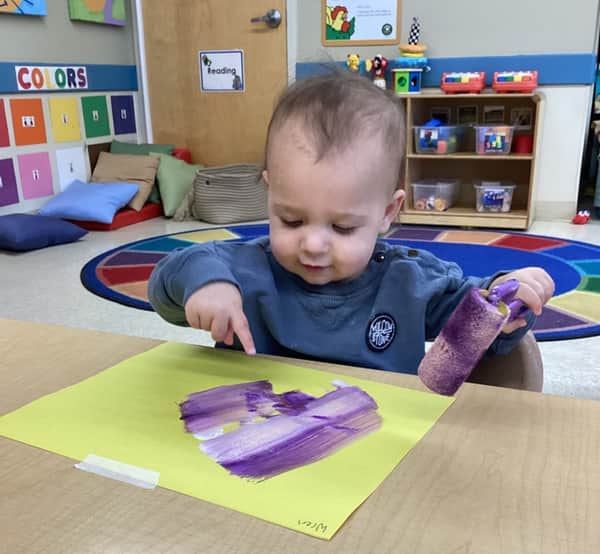 Young toddler rolling purple paint across yellow paper during an art activity at Primrose School of Thornton