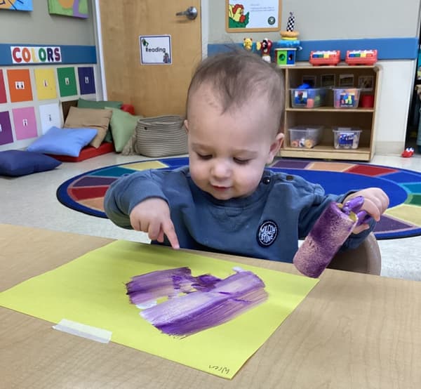Young toddler rolling purple paint across yellow paper during an art activity at Primrose School of Thornton