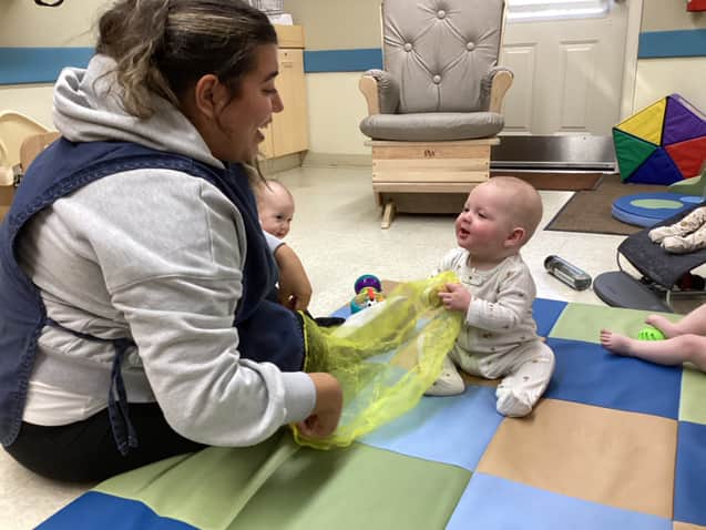 Infant teacher playing with a yellow scarf alongside two babies on a mat in the Infant Room