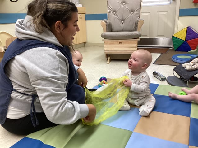 Infant teacher playing with a yellow scarf alongside two babies on a mat in the Infant Room