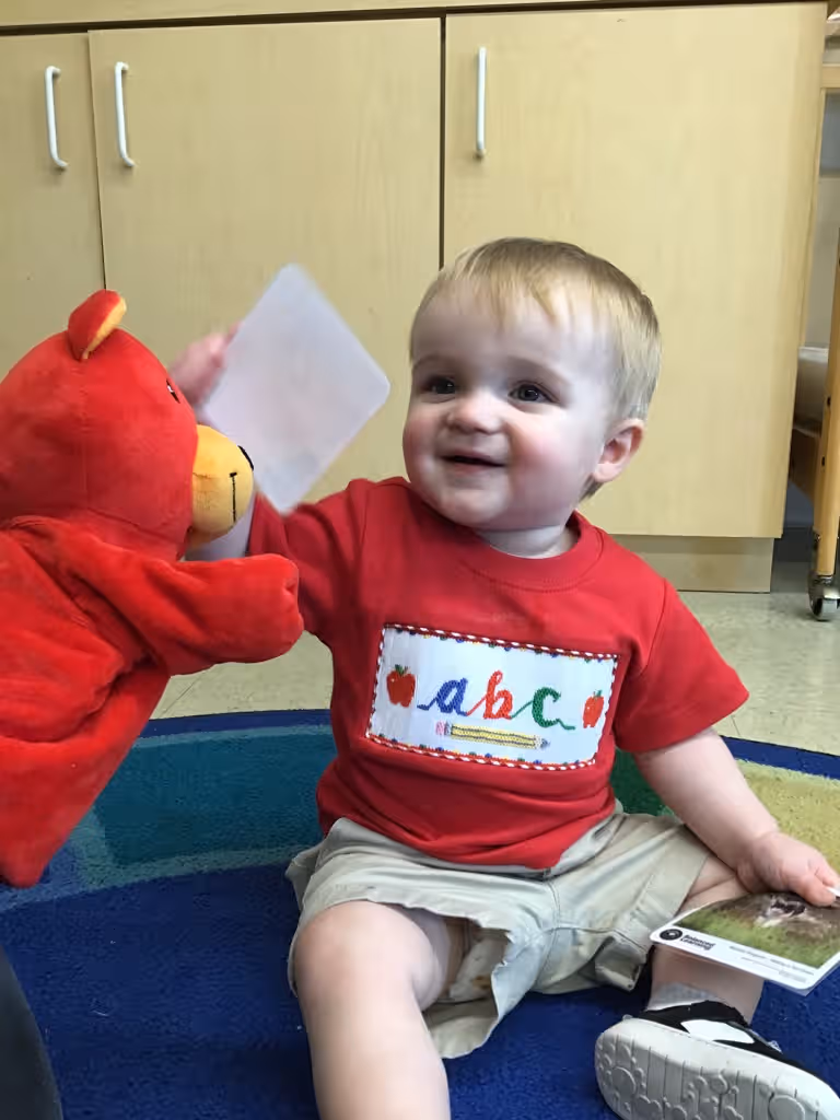 Toddler child wearing a red ABC shirt smiling holding a flash card with Benjamin the bear, a red stuffed bear puppet