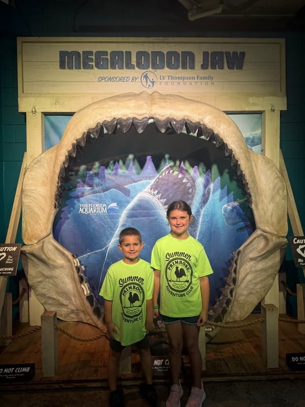 Two children posing inside a giant megalodon jaw exhibit at an aquarium while participating in a summer adventure learning excursion.