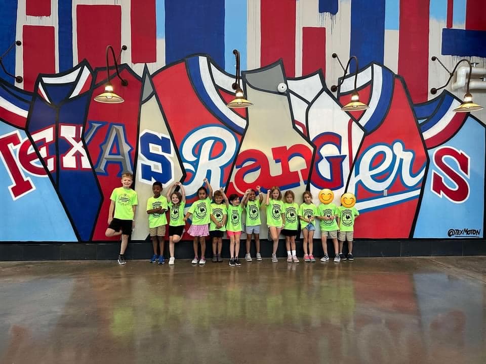 Group of Primrose School summer campers in bright shirts posing in front of a colorful Texas Rangers mural during a field trip to the stadium, celebrating sports, teamwork, and local culture.