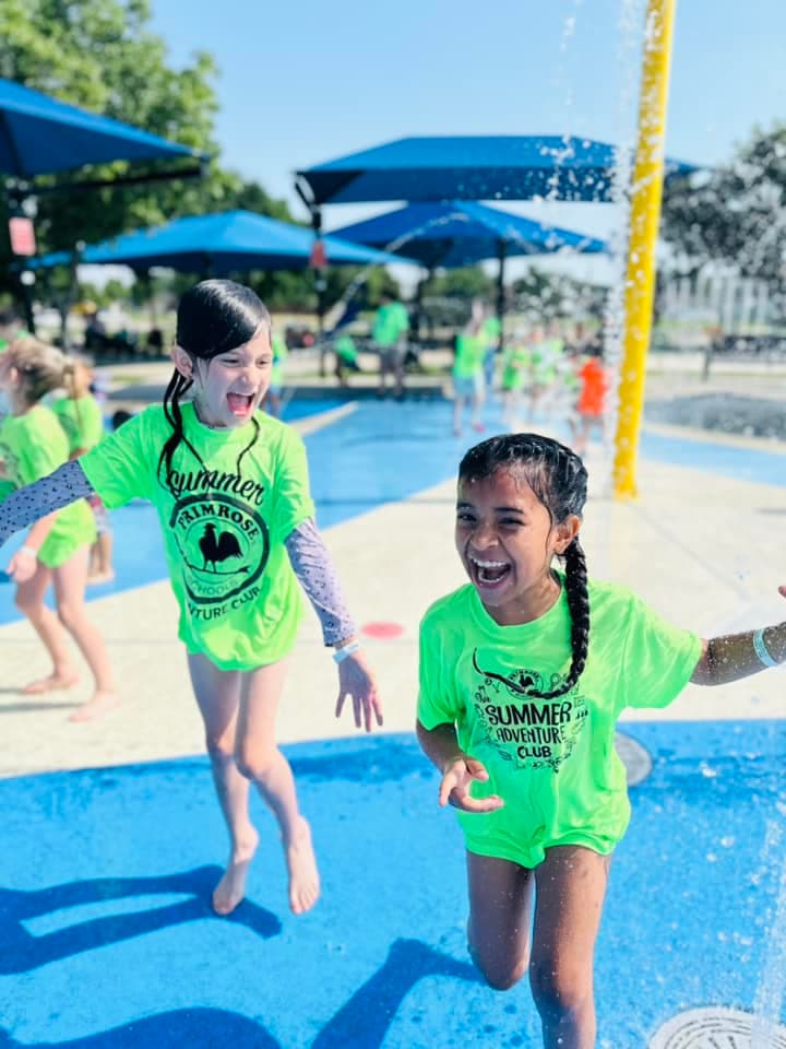 Primrose School at Heritage summer camp students laughing and playing at a splash pad in bright green shirts under blue umbrellas in Fort Worth, TX