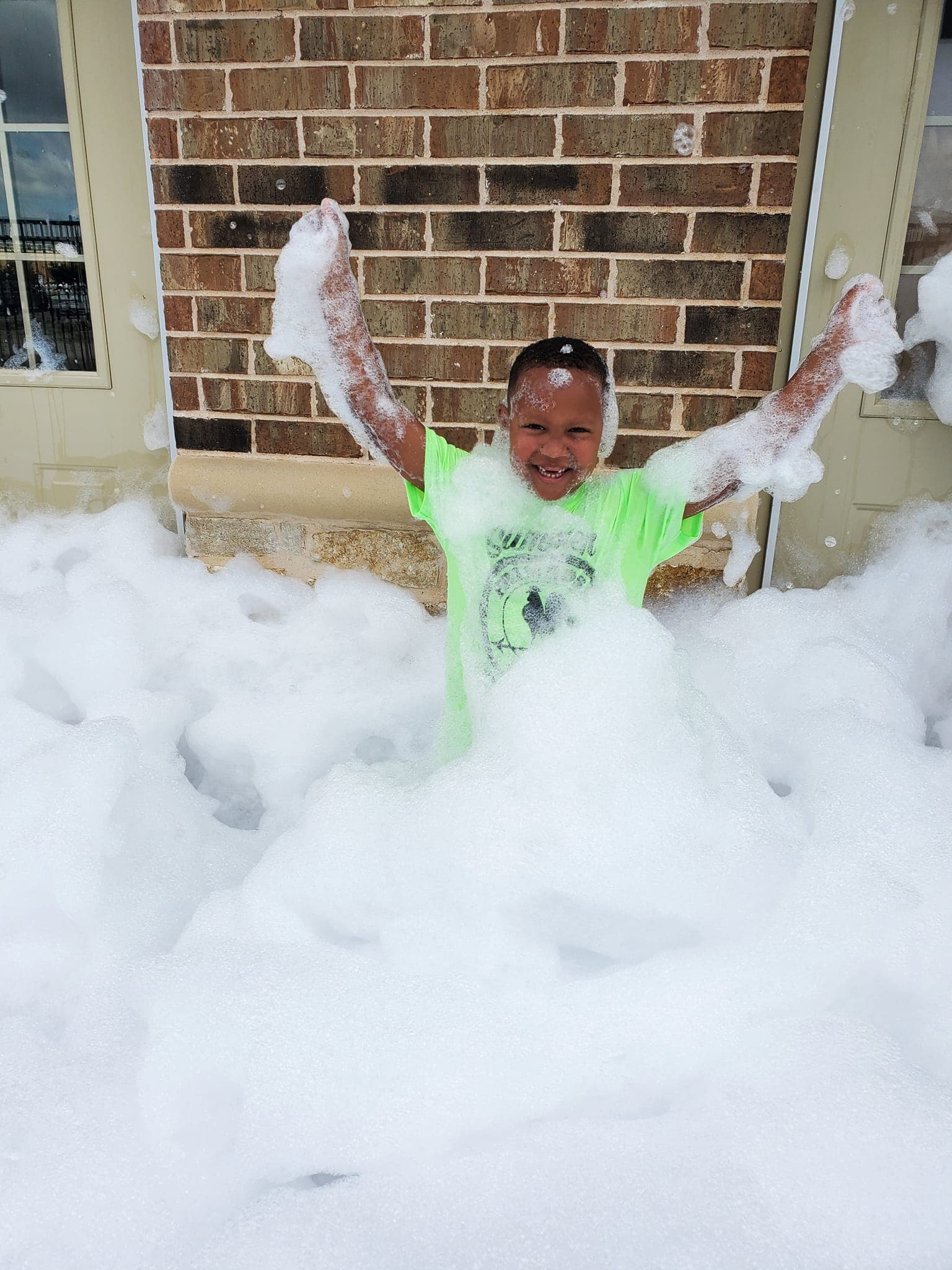 Primrose School summer camper playing in thick white foam outdoors during a Foam Day activity, celebrating summer fun and sensory play with big smiles and laughter at the best summer camp in Haslet.