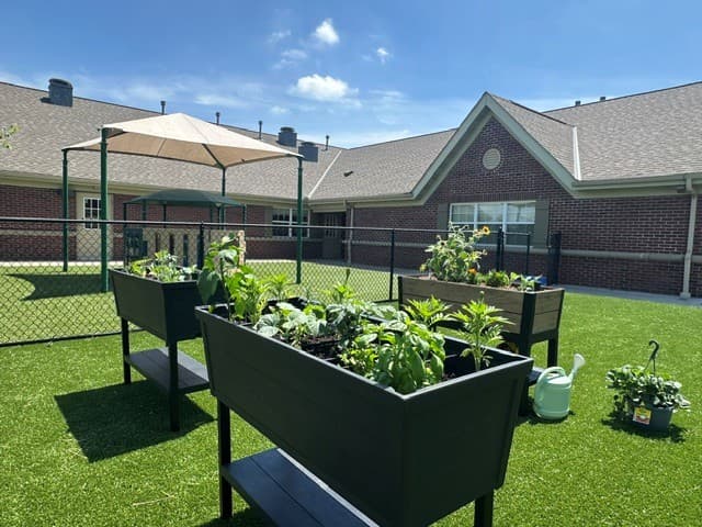 The Primrose Patch raised garden beds at Primrose School of Eagan featuring lush vegetables and herbs growing on artificial turf in the school courtyard with the brick building and shade sail in the background