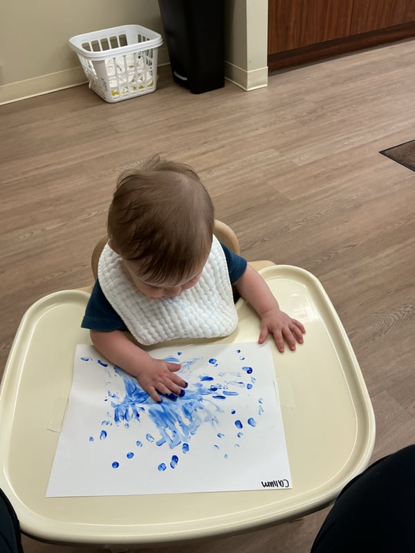 An infant sitting in a highchair fingerpainting with blue paint.