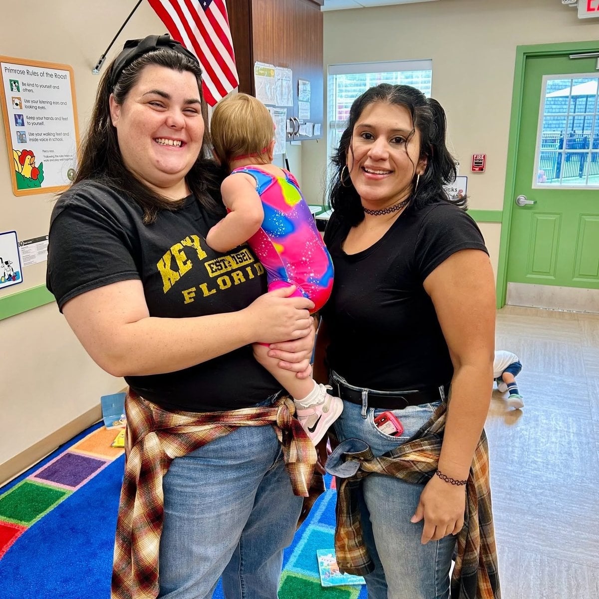 Two infant care teachers smiling with a baby in the classroom at Primrose School of Woburn, MA