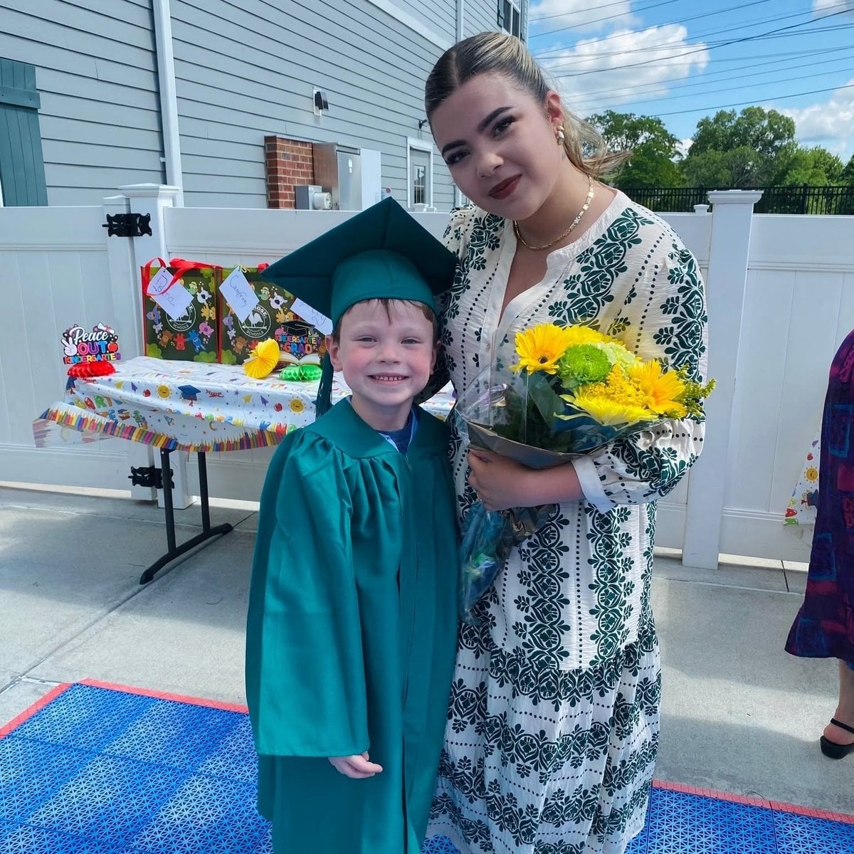 Kindergarten graduate in cap and gown with family member at Primrose School of Woburn, MA graduation ceremony
