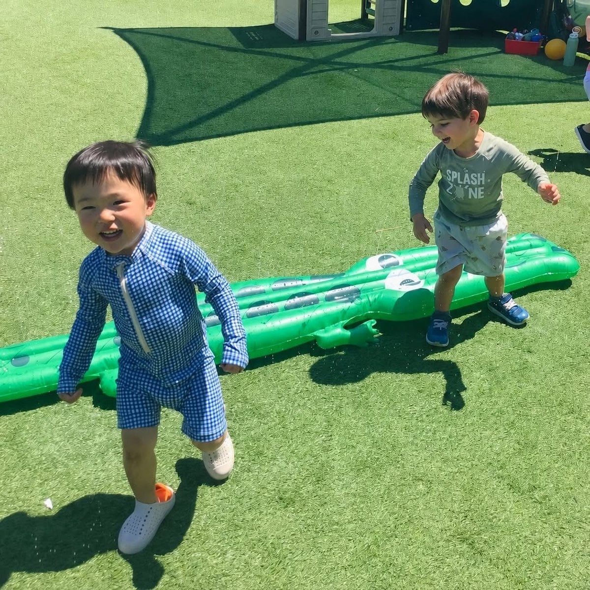 Preschool children laughing and playing outdoors on a water inflatable at Primrose School of Woburn, MA