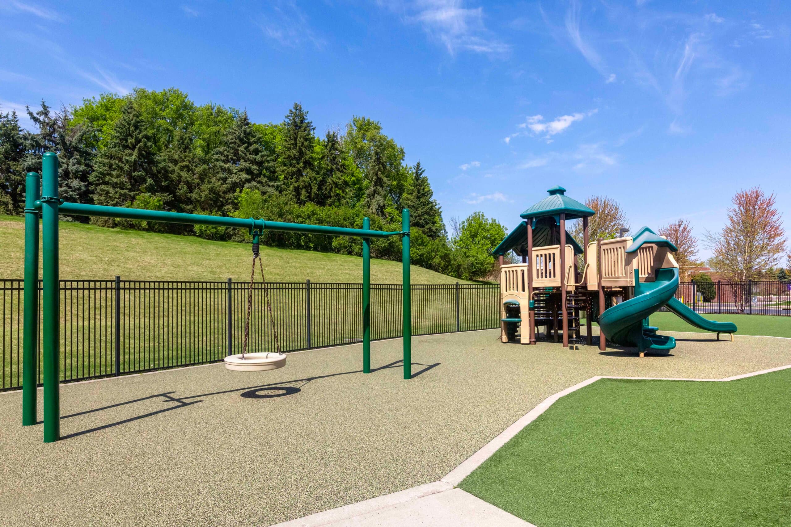 Pre-Kindergarten outdoor play area at Primrose School of Eagan featuring a multi-level climbing structure with spiral slide, disc swing, artificial turf, and rubber safety surfacing with a natural wooded hillside backdrop
