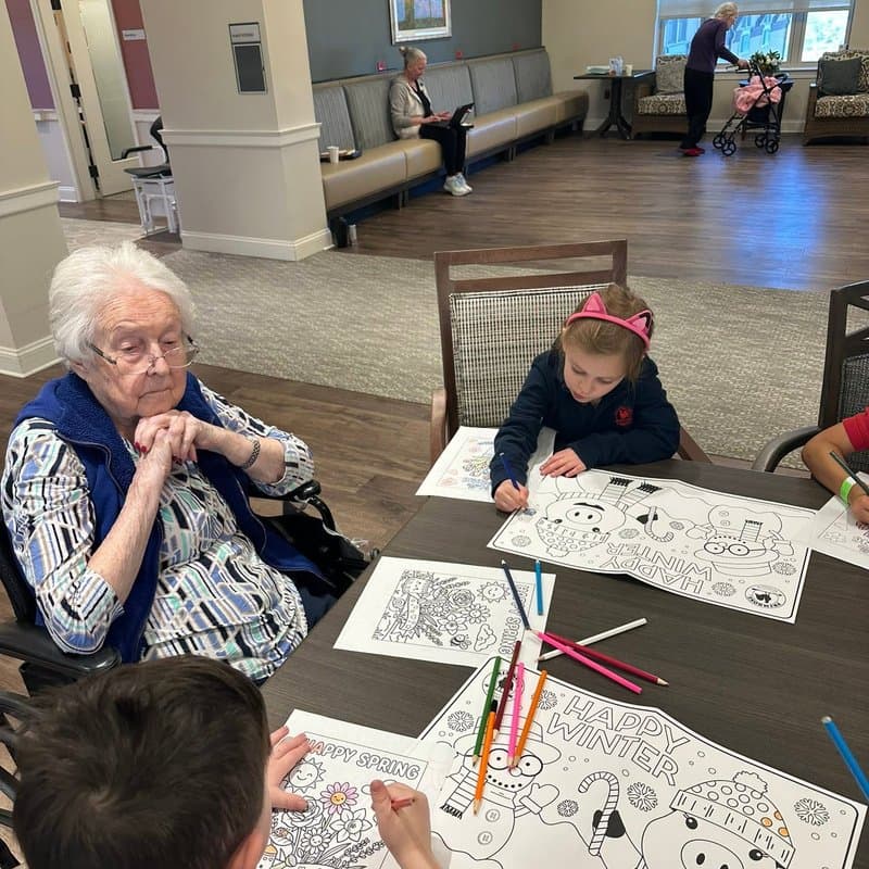 Children coloring seasonal activity sheets alongside older adults during a creative learning activity associated with Primrose School at Highlands Ranch Business Park.