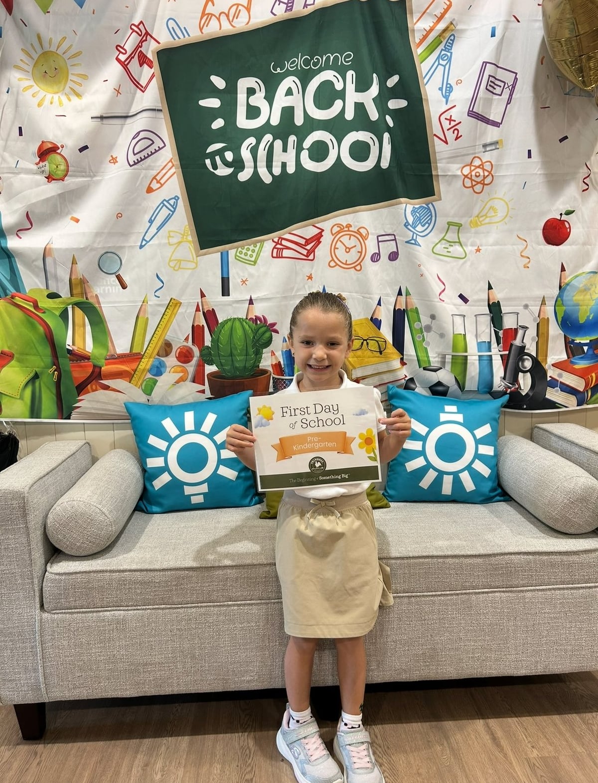 girl in khaki skirt and white shirt holding a first day of prekindergarten sign in front of gray couch and back to school backdrop