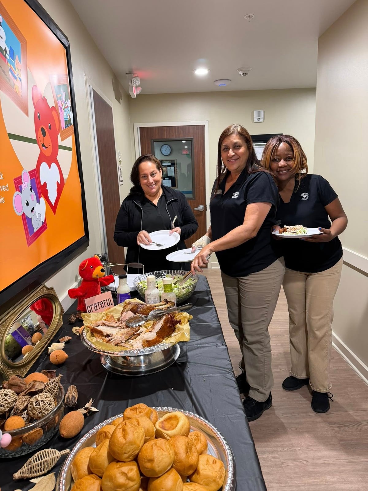 Three adult women in black polos and khaki pants holding plates of food standing in front of a table of food