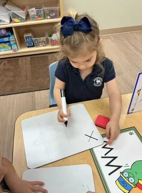 girl in blue dress holding a marker writing the letter x on a white board on a table