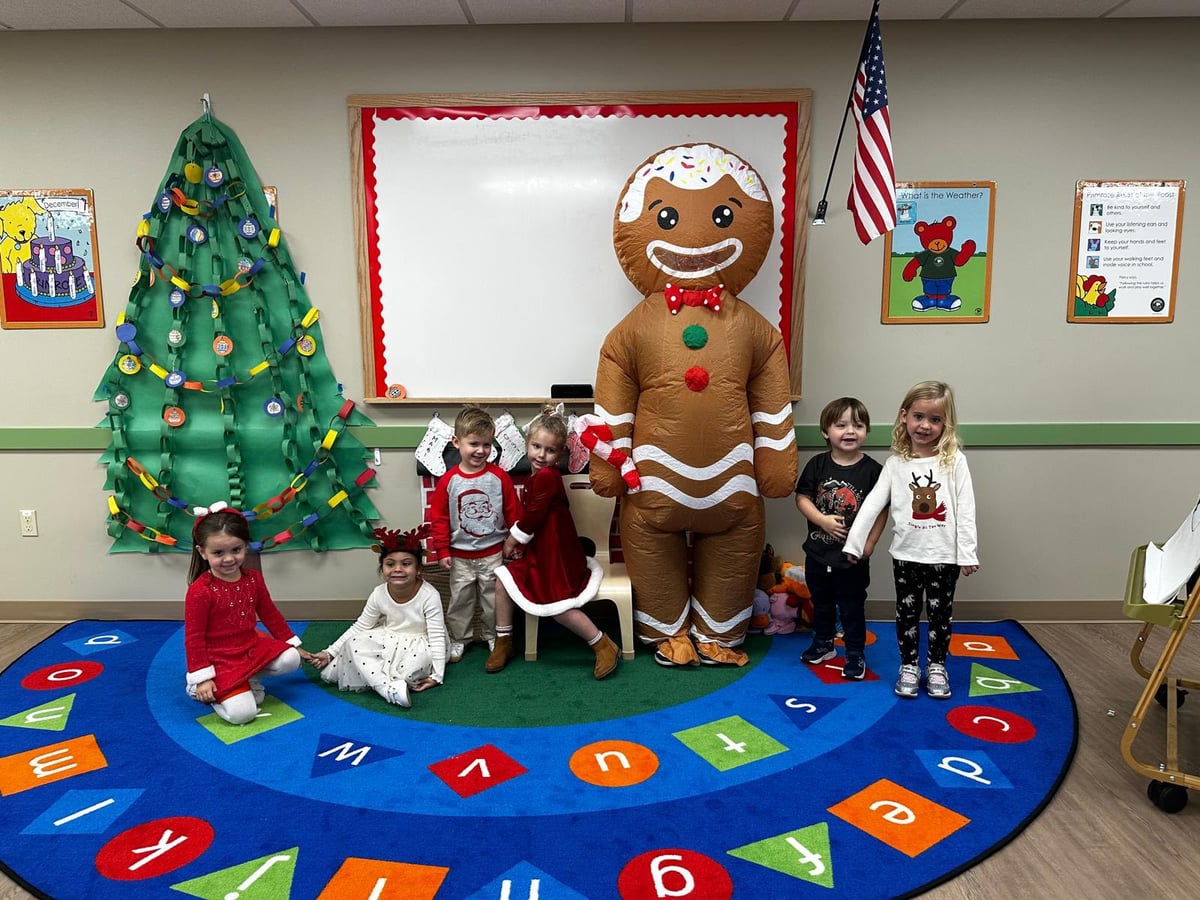 Six children dressed in holiday outfits standing with a giant blow up gingerbread. There is a paper Christmas tree decoration on the wall and a blue rug.