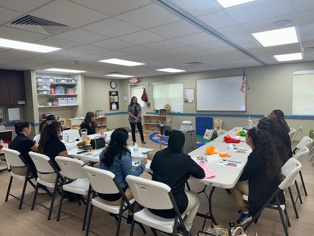 women sitting in white chairs in a classroom with tables and puppets in front of them