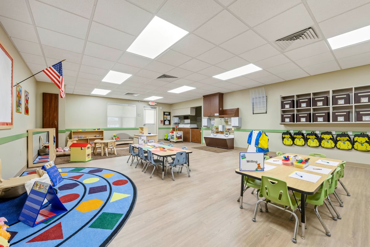 classroom with blue rug that has shapes on it two tables one with green chairs one with blue and cubbies on the wall