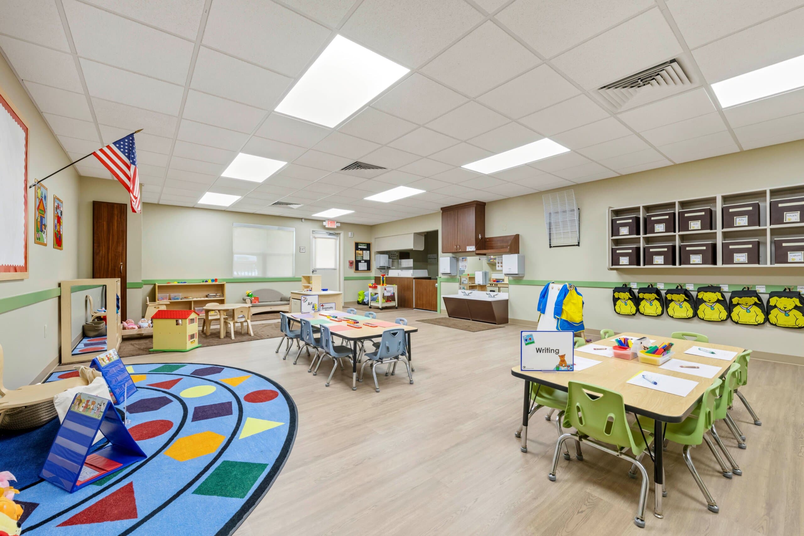 classroom with blue rug that has shapes on it two tables one with green chairs one with blue and cubbies on the wall