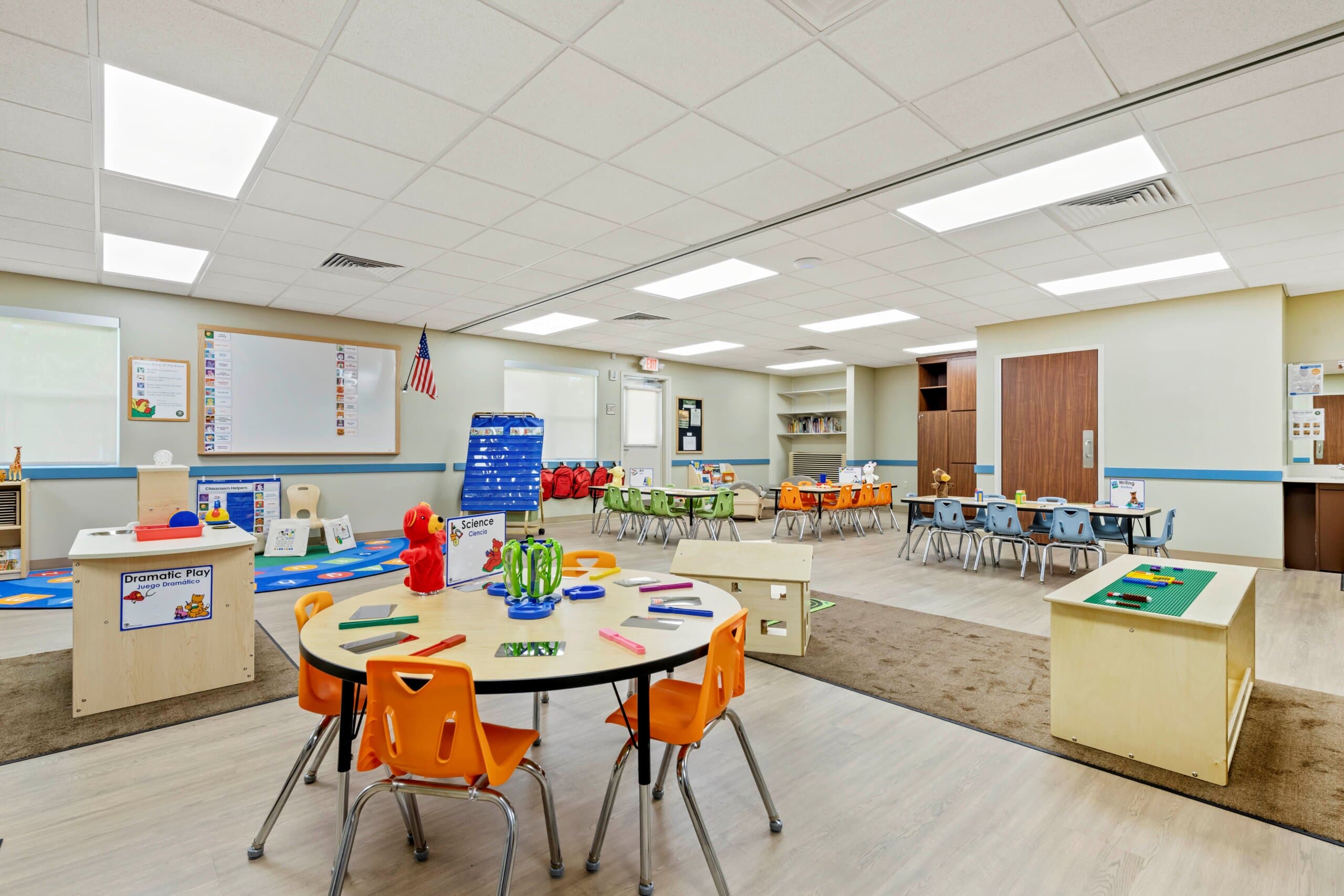 classroom with circle table and orange chairs, lego table, three rectangular tables with blue chairs orange chairs and green chairs, teacher easel with blue attendance chart and white board with American Flag hanging next to it