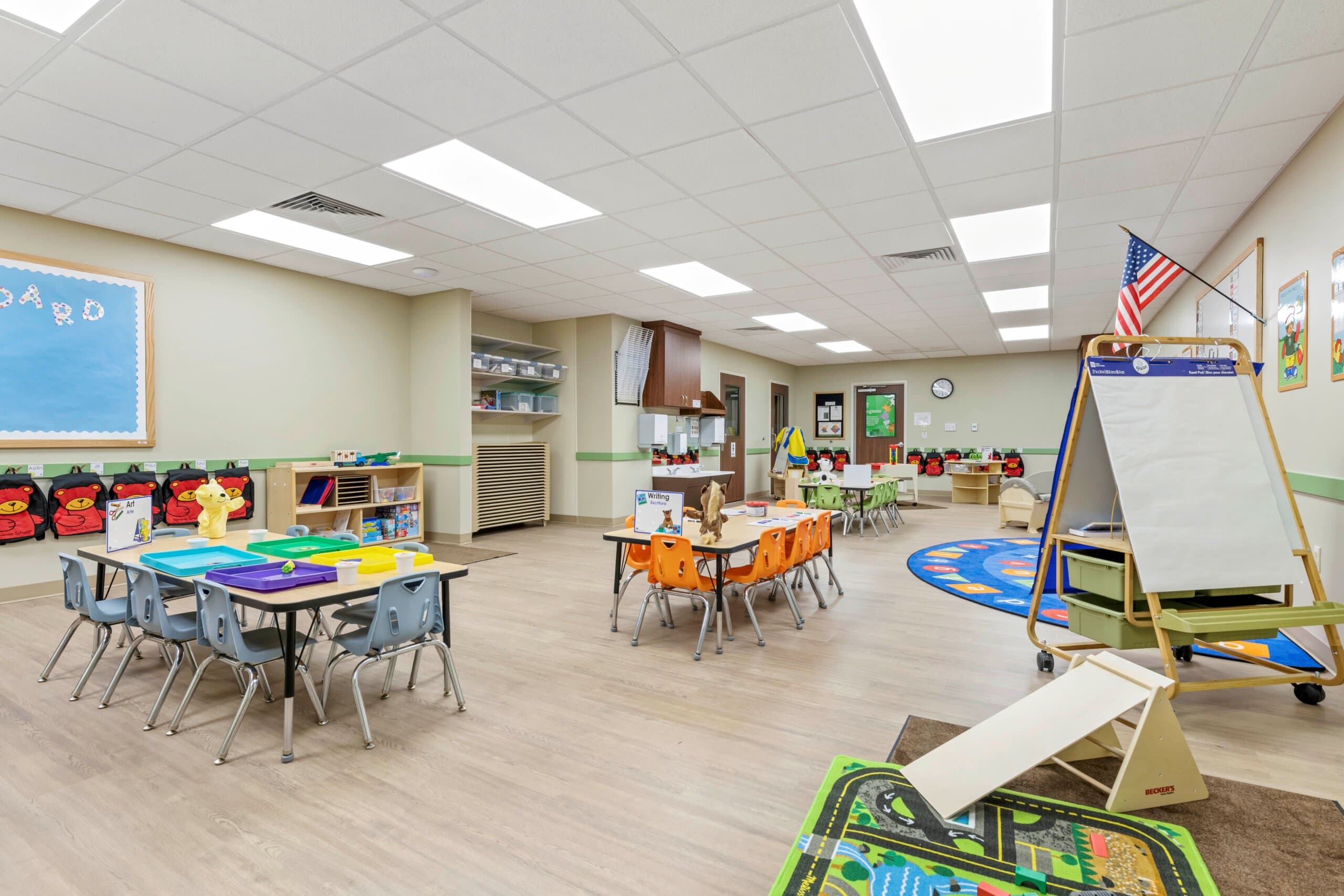 Classroom with three tables blue orange and green chairs, easel with paper, American flag, ramp on car rug and backpacks hanging with red bear