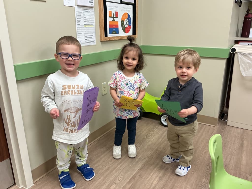 Three Young Primrose School of Forest Hill children sharing and displaying artwork in a Early Preschool classroom setting