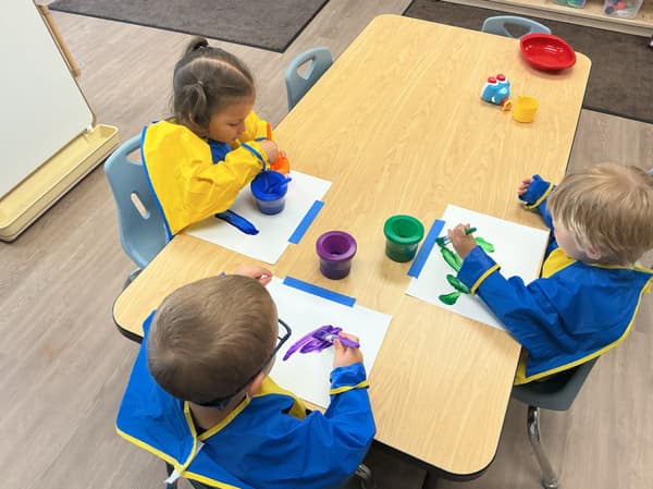 Three early preschool children at Primrose School of Forest Hill sitting together at a table, using paints and art materials to explore creativity.