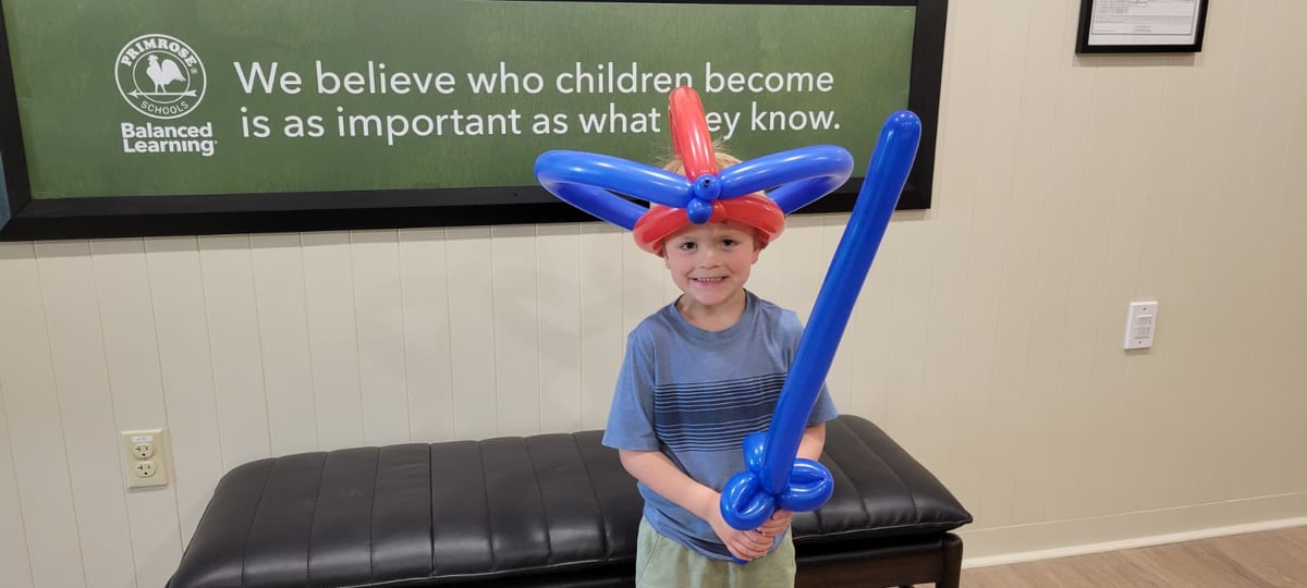 Preschool student at Primrose School of Forest Hill smiling while wearing a colorful balloon hat and holding a balloon sword after a special balloon entertainer visit, celebrating creativity, imagination, and joyful learning.