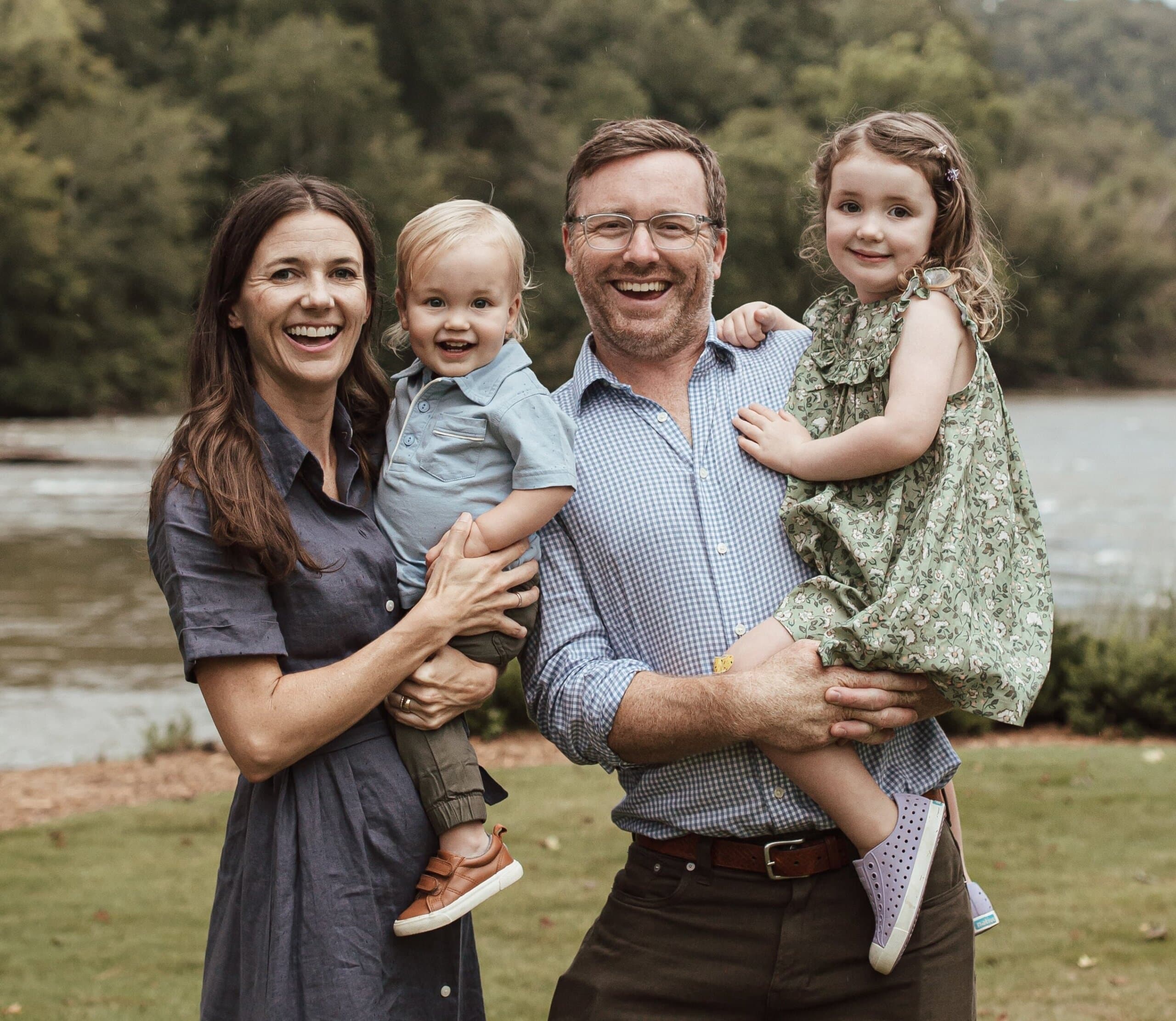 A smiling family of four standing outdoors by a river with wooded hills in the background. The mother, wearing a blue dress and brown sandals, is holding a young blond boy in a light blue shirt. The father, in a checkered shirt and dark pants, is holding a little girl in a green dress. The family looks happy and relaxed together on the grassy riverbank.